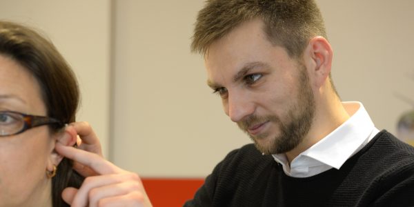 Ein Auszubildender zum Hörakustiker befestigt ein Hörgerät am Ohr einer Kundin.