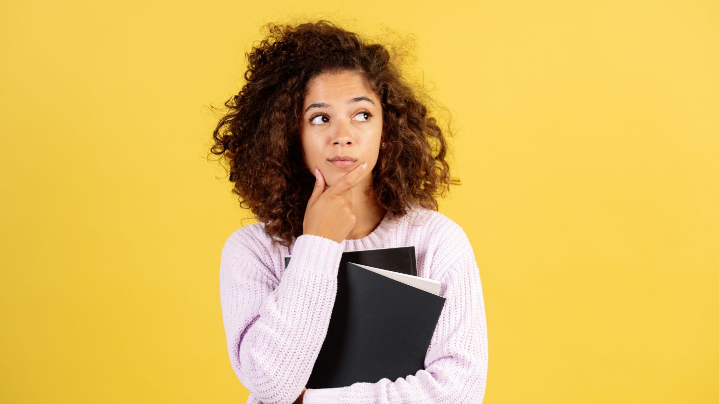 junge Frau steht vor einem gelben Hintergrund und guckt nachdenklich in die Luft