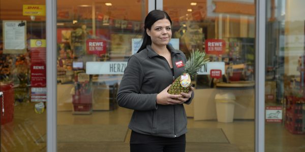 Eine Auszubildende zur Einzelhandelskauffrau steht vor dem Supermarkt und hält eine Ananas in der Hand.