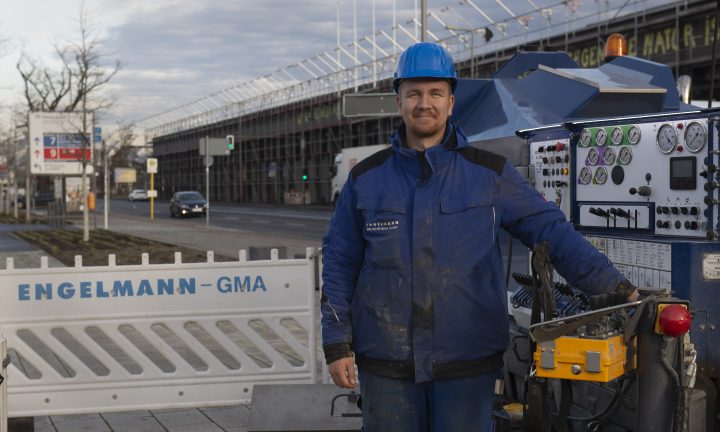 Ein Auszubildender zum Brunnenbauer lächelt in blauer Arbeitskleidung in die Kamera.