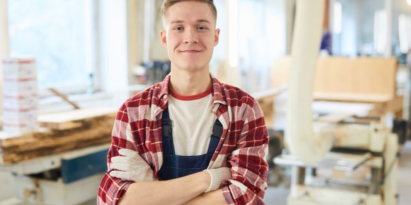 Ein Auszubildender zum Zimmerer steht mit Handschuhen und Arbeitskleidung in seiner Werkstatt.