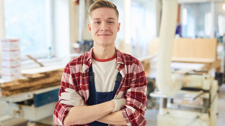 Ein Auszubildender zum Zimmerer steht mit Handschuhen und Arbeitskleidung in seiner Werkstatt.