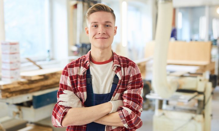Ein Auszubildender zum Zimmerer steht mit Handschuhen und Arbeitskleidung in seiner Werkstatt.