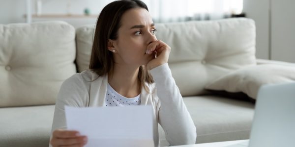 Eine junge Frau hält zu Hause am Schreibtisch eine Rechnung in der Hand und denkt an finanzielle Probleme.