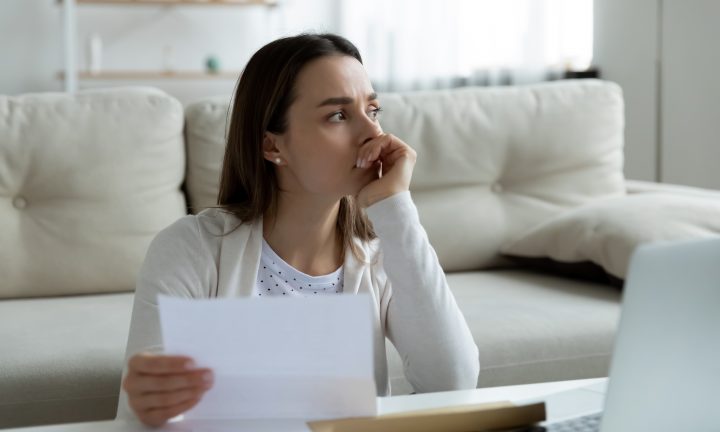 Eine junge Frau hält zu Hause am Schreibtisch eine Rechnung in der Hand und denkt an finanzielle Probleme.