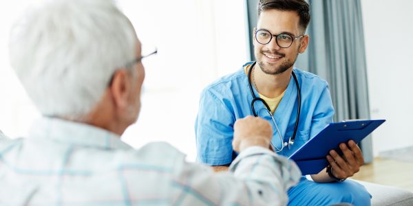 Ein Auszubildender zum Pflegefachmann sitzt im Pflegeheim auf einem Sofa und unterhält sich mit einem älteren Herrn.