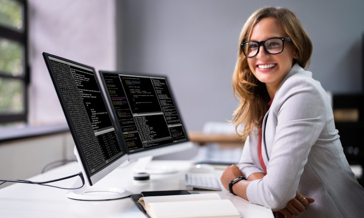 Eine Auszubildende im IT-Bereich sitzt an ihrem Schreibtisch, auf dem zwei Monitore mit Programmiersprache stehen.