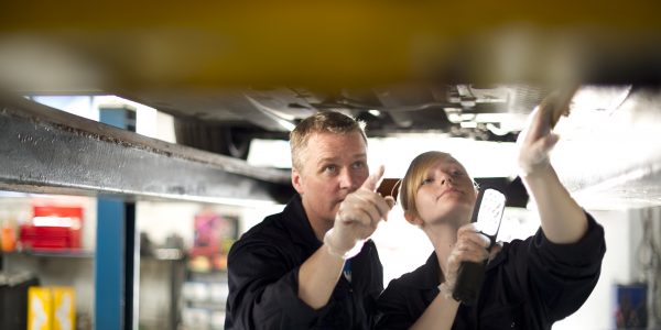 ein Ausbilder und eine Auszubildende arbeiten an einem Auto, dass auf einer Hebebühne steht