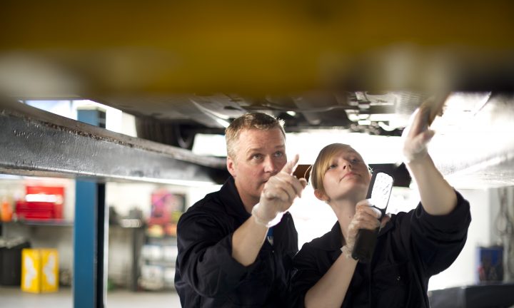 ein Ausbilder und eine Auszubildende arbeiten an einem Auto, dass auf einer Hebebühne steht