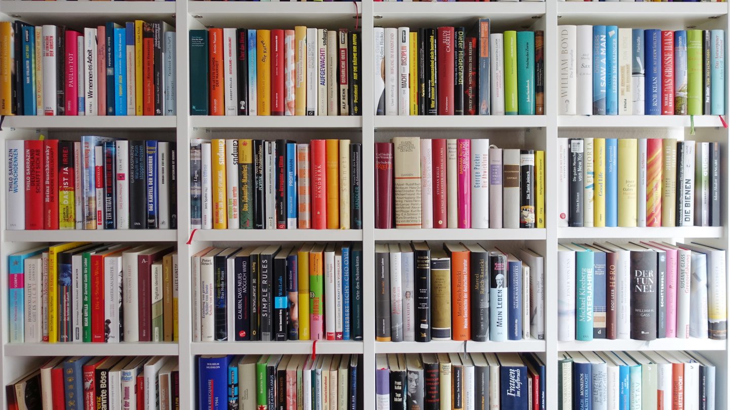 Bunte Bücher stehen eng gedrängt in einem weißen Bücherregal.