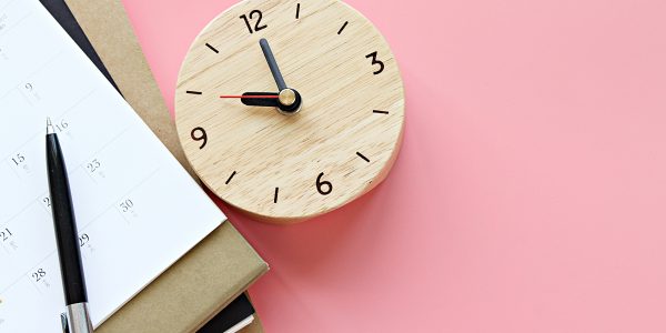 Auf einem Tisch liegen ein Kalender, Notizbücher, ein Stift und eine Uhr.