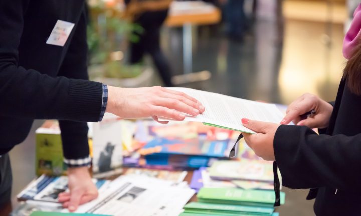 Ein Mann und eine Frau reichen ein Informationsblatt über den Messestand.