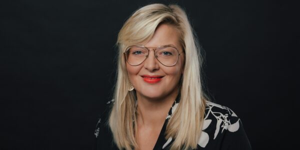 Blonde Frau mit rotem Lippenschrift und goldener Brille lächelt in Kamera