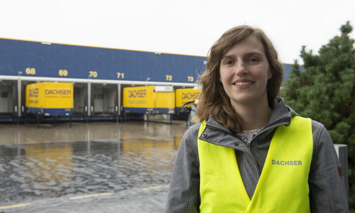 Kauffrau für Spedition und Logistikleistungen steht mit gelber Weste vor Lagerhallen.