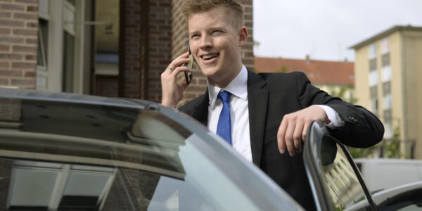 Auszubildender zum Fachwirt für Versicherungen und Finanzen telefoniert lachend und steht am Auto.