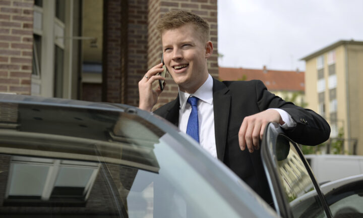 Auszubildender zum Fachwirt für Versicherungen und Finanzen telefoniert lachend und steht am Auto.