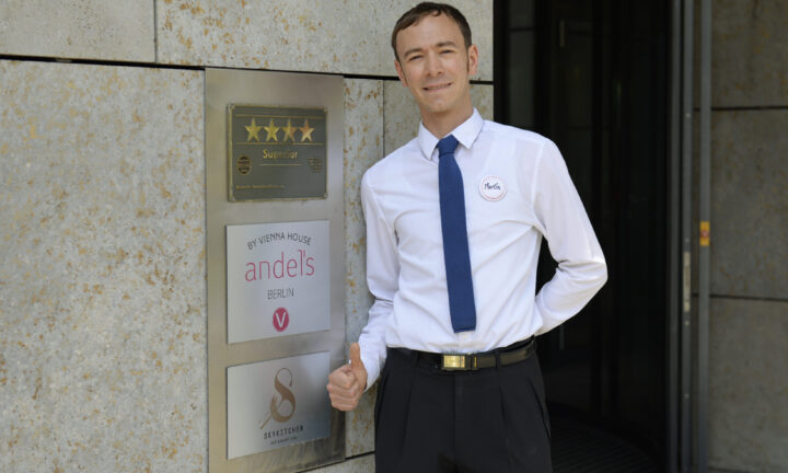 Hotelfachmann steht vor den Türen des Vienna House Andel’s Berlin und zeigt auf Auszeichnungen.
