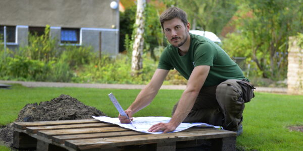 Junger Gärtner hält einen Zollstock und hat einen großen Plan vor sich ausgebreitet.
