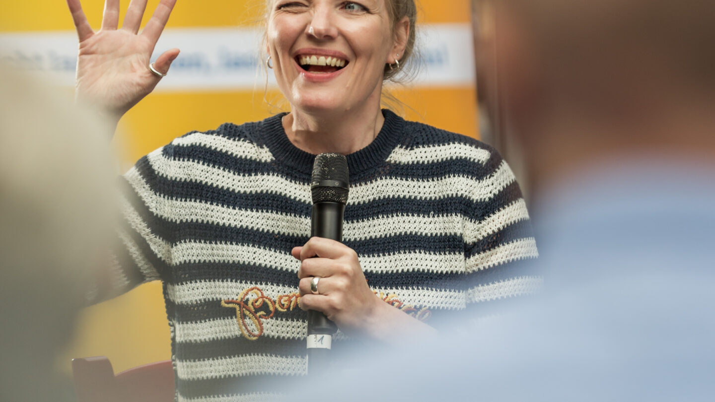 Moderatorin kneift das rechte Auge zusammen und hält die rechte Hand hoch