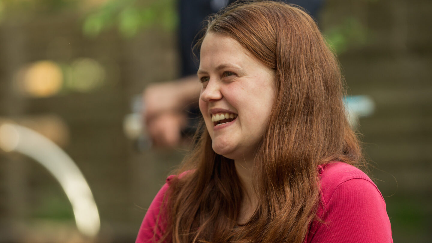 eine junge Frau sitzt auf einem Stuhl und lacht. Im Hintergrund steht ein Mann