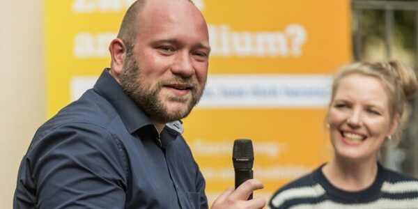 Ein junger Mann mit Mikrofon lacht. Dahinter sitzt eine Frau, die ihn anlacht.