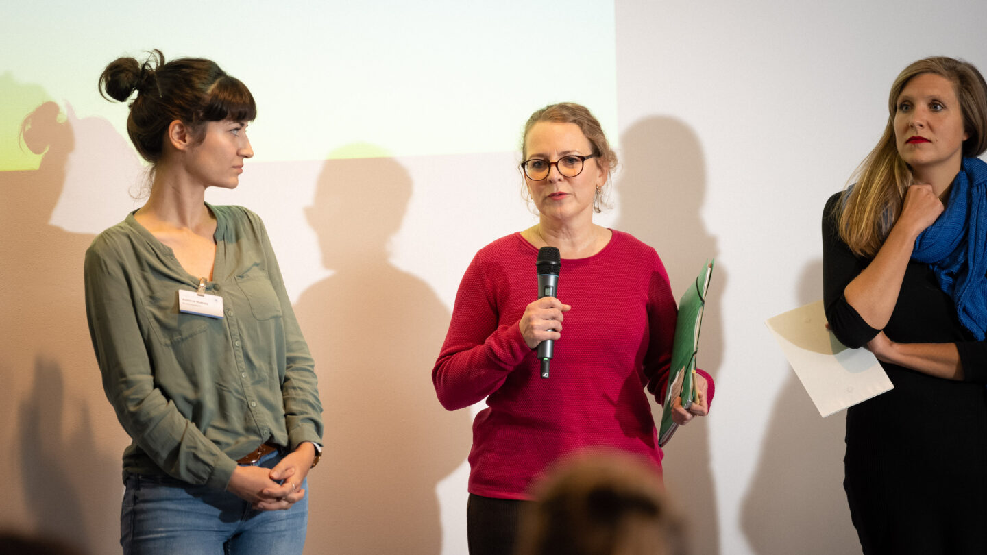 Drei Frauen stehen auf der Bühne. Die Frau in der Mitte spricht in das Mikrofon