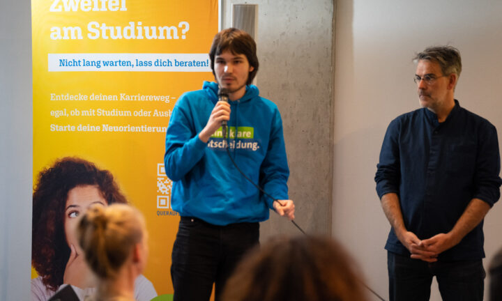 Ein junger Mann spricht ins Mikrofon. Rechts daneben steht ein mittelalter Mann und hört zu