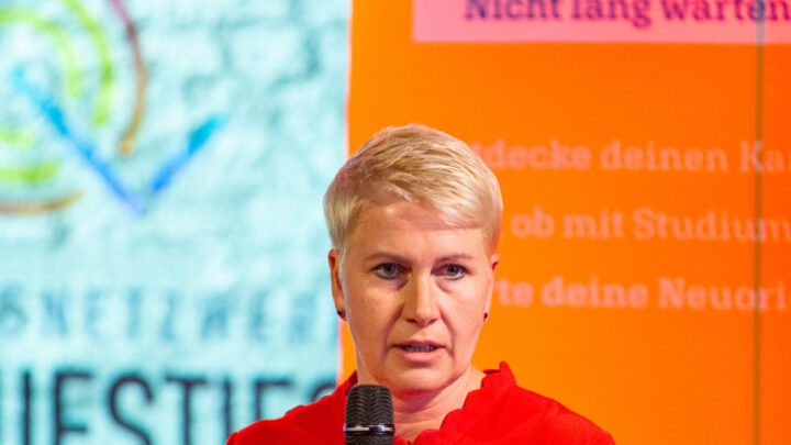 Eine Frau mit blonden Haaren und rotem Pullover erzählt. Sie hat ein Mikrofon in der Hand.