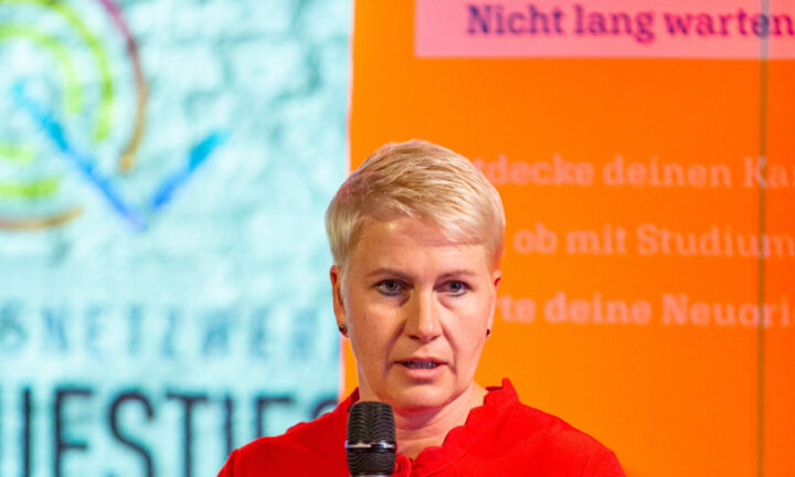 Eine Frau mit blonden Haaren und rotem Pullover erzählt. Sie hat ein Mikrofon in der Hand.