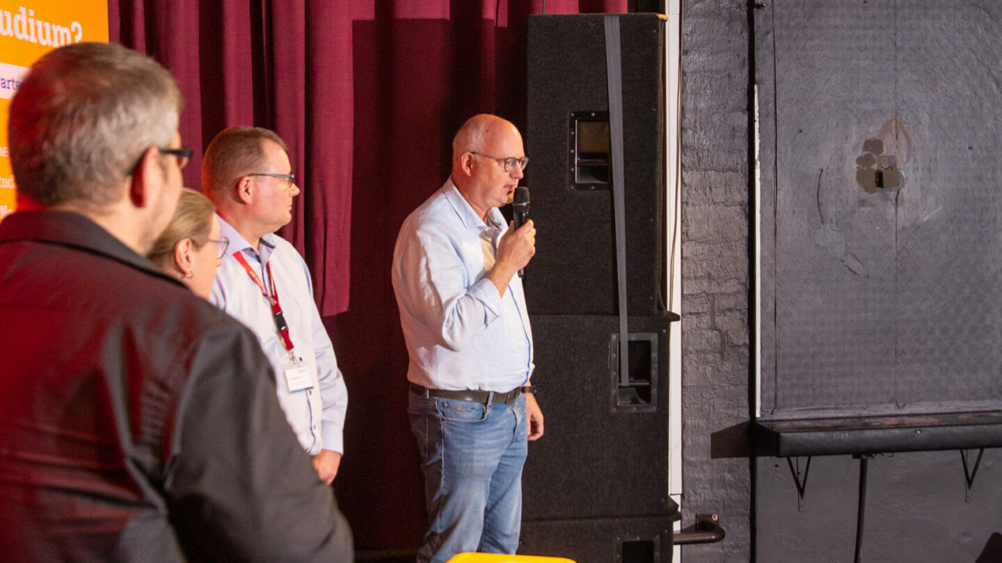 Vier Personen stehen auf der Bühne. Ein Mann spricht in das Mikrofon.