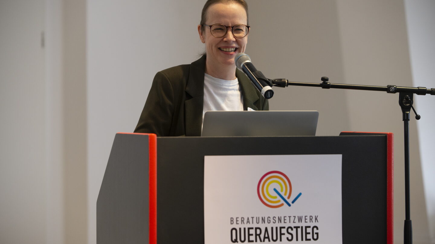 Eine Frau mit runder Brille steht am Rednerpult und lächelt in die Kamera