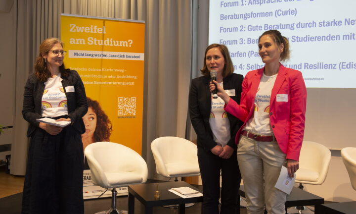 Auf einer Bühne reicht eine Frau einer anderen Frau das Mikrofon zum Sprechen, während eine weitere Frau die beiden beobachtet