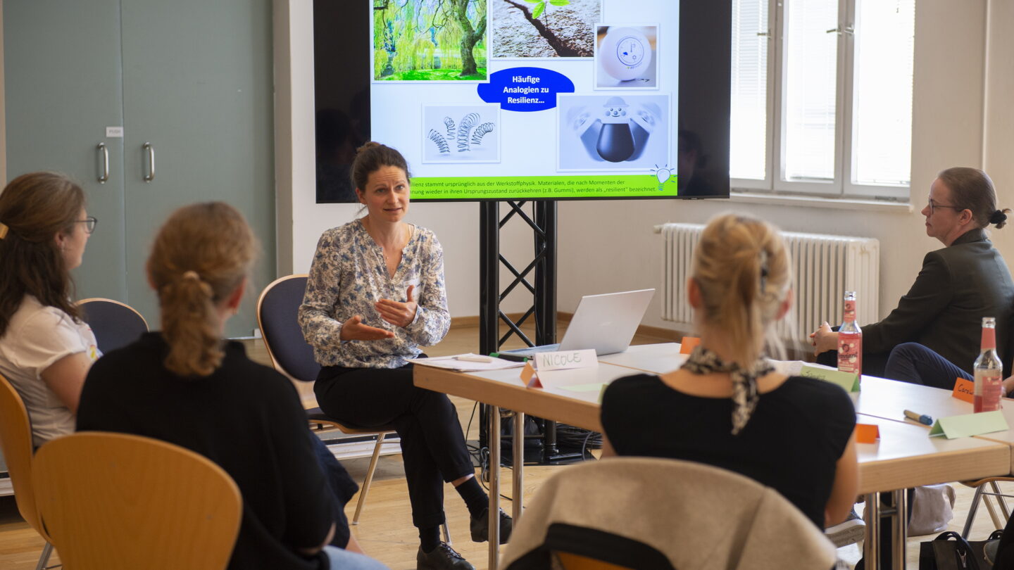Um einen Tisch herum sitzen mehrere Personen und richten ihren Blick auf die Rednerin