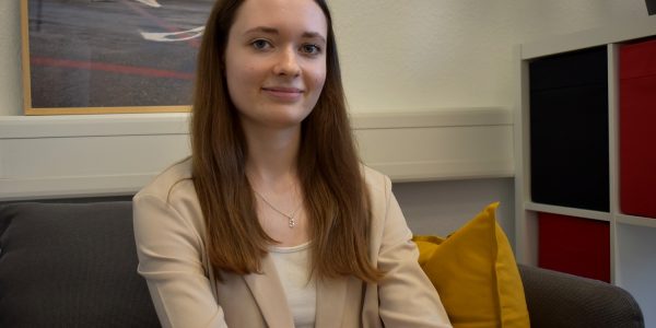 Eine junge Frau mit langen dunkelblonden Haaren sitzt in einem Sessel vor einem Bild und einem Regal und lächelt freundlich