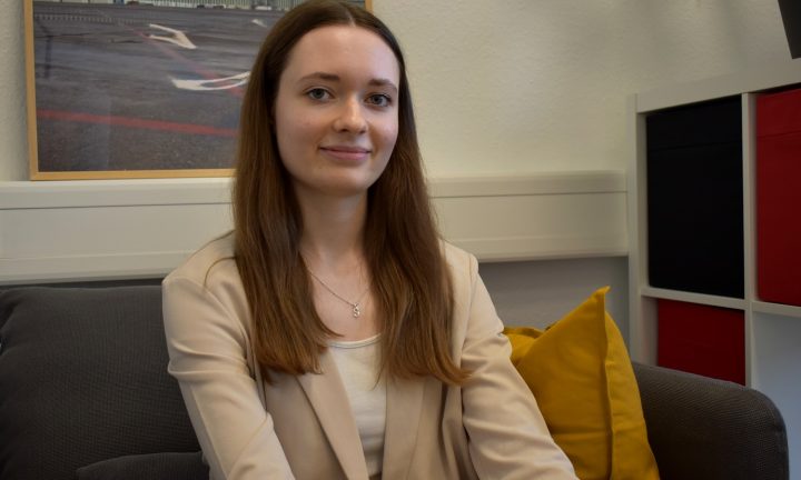 Eine junge Frau mit langen dunkelblonden Haaren sitzt in einem Sessel vor einem Bild und einem Regal und lächelt freundlich