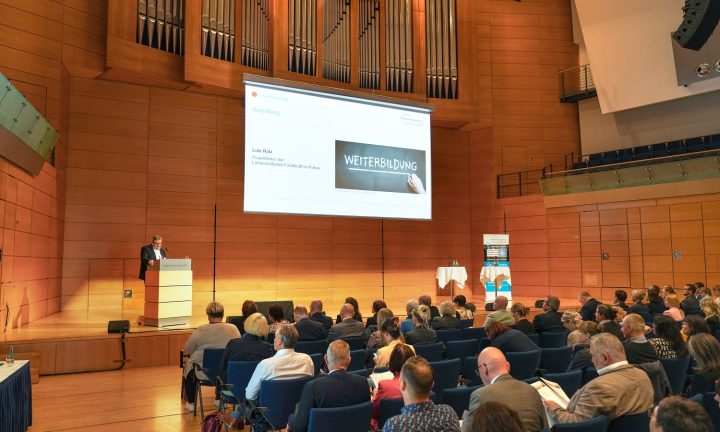 Viele Menschen sitzen in einem großen Veranstaltungssaal und lauschen den Ausführungen eines Referenten zum Thema Weiterbildung