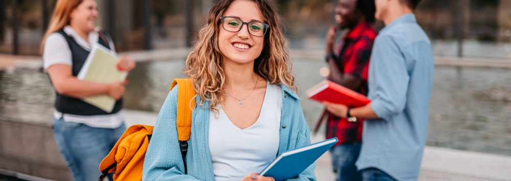 Eine lächelnde Studentin steht mit ihrem Rucksack und einem Buch auf dem Campus. Im Hintergrund unterhalten sich ihre Freunde miteinander.