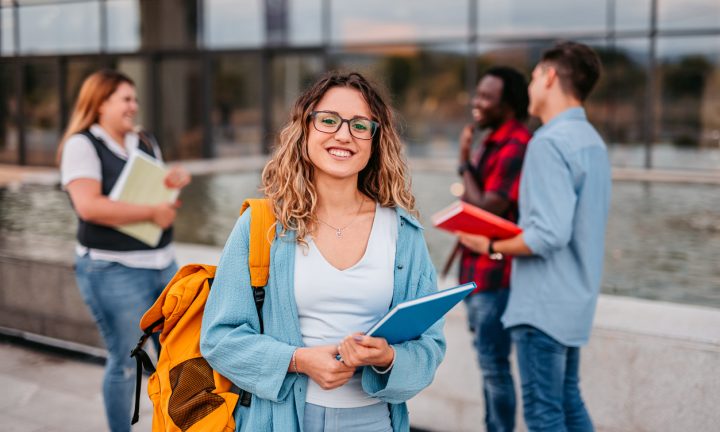 Eine lächelnde Studentin steht mit ihrem Rucksack und einem Buch auf dem Campus. Im Hintergrund unterhalten sich ihre Freunde miteinander.
