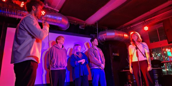 Drei Poetry-Slammer*innen stehen zusammen mit zwei Moderator*innen auf einer rosarot beleuchteten Bühne.