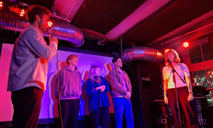 Drei Poetry-Slammer*innen stehen zusammen mit zwei Moderator*innen auf einer rosarot beleuchteten Bühne.