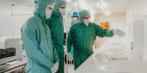 Drei Mikrotechnologen tragen Schutzkleidung und unterhalten sich in einem Reinraum.