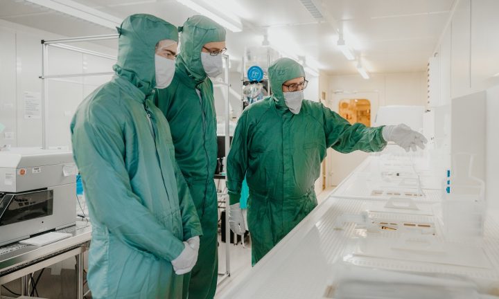 Drei Mikrotechnologen tragen Schutzkleidung und unterhalten sich in einem Reinraum.