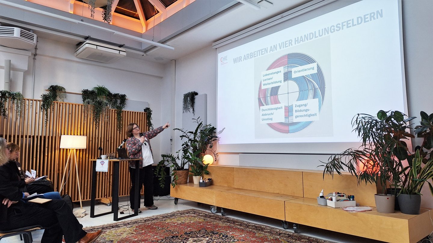 Eine Frau steht auf einer Bühne mit vielen Pflanzen und zeigt auf eine Präsentationsfolie.