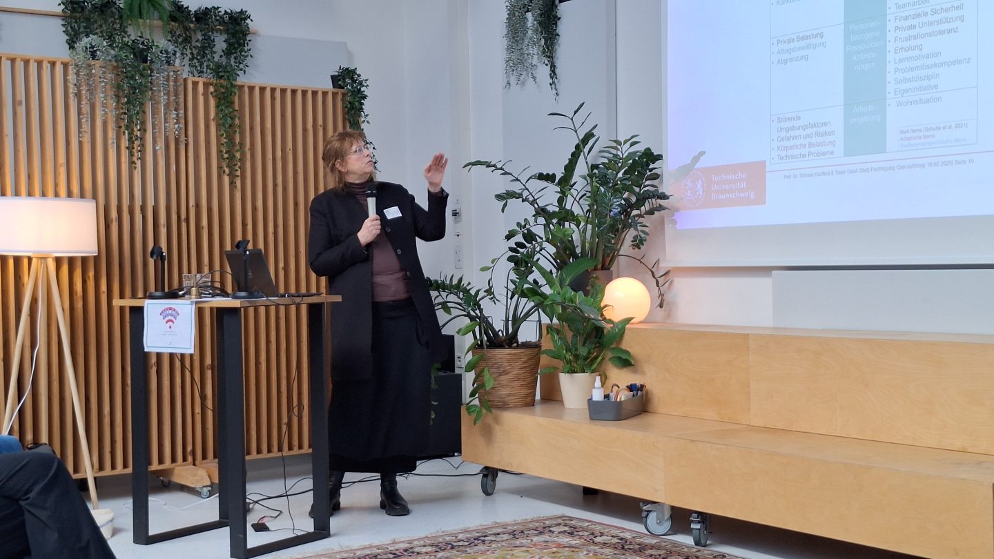 Eine Frau steht am Rednerpult mit Pflanzen im Hintergrund und zeigt auf eine Präsentationsfolie.