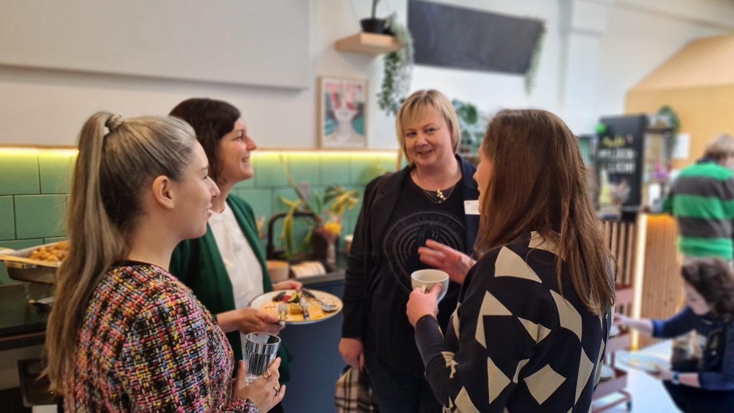 Vier Frauen sprechen auf einer Tagung miteinander und halten Getränke und Teller in den Händen.