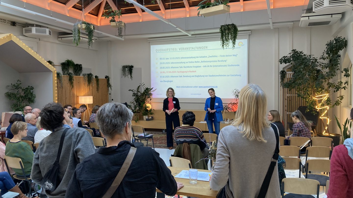 Zwei Moderatorinnen lächeln und sprechen mit dem Publikum, das sitzt bzw. an Stehtischen steht.