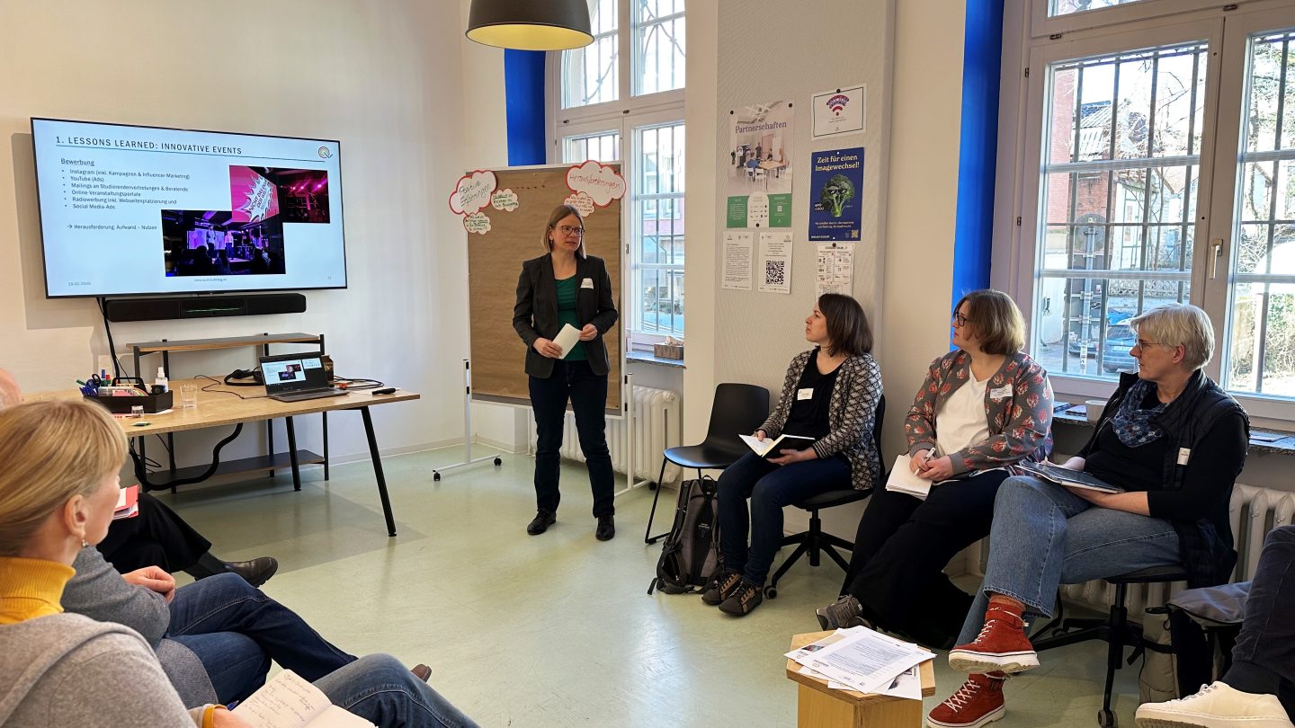Eine Frau präsentiert auf einem Bildschirm ihren Input vor einem kleinen Publikum, das im Halbkreis um sie herumsitzt.