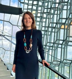 Eine Frau mit langen hellbraunen Haaren steht auf einer Treppe in einem Gebäude mit gläsernen Wänden.