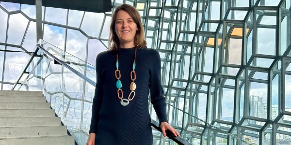 Eine Frau mit langen hellbraunen Haaren steht auf einer Treppe in einem Gebäude mit gläsernen Wänden.